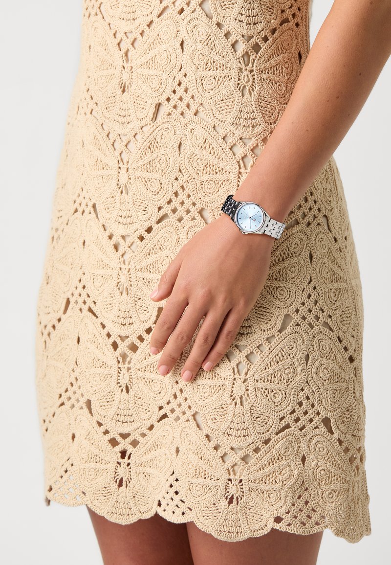 Silver watch with blue dial and white-black patterned strap worn over a crochet dress featuring butterfly motifs in beige.