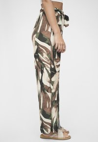 Mønstrete, lettvekts bukser med snor i livet, med abstrakte bladdesign i grønt, brunt og kremfarge. Synlige flate sandaler.