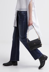 Bolso negro con cadena, combinado con una blusa blanca y un cárdigan largo. Unos jeans de mezclilla oscuros y botas de tobillo completan el atuendo.