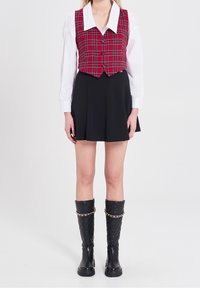 Czerwono-zielona kratkowana kamizelka na krótko, noszona na białej, długiej koszuli, w zestawie z czarną plisowaną spódnicą i czarnymi, wysokimi, pikowanymi butami do kolan z łańcuchem.