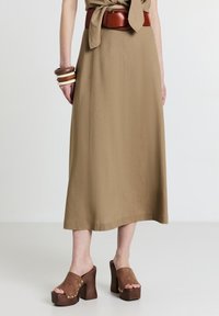 Abito midi marrone con dettaglio in vita legato, abbinato a zoccoli marroni con tacco decorati con finiture dorate e bracciali in legno impilati.