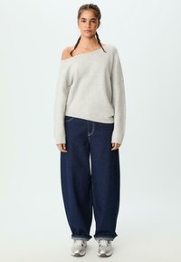 Helles graues Off-Shoulder-Pullover mit gerippten Bündchen, kombiniert mit lockeren, dunkelblauen Jeans aus Denim, die über einen kontrastierenden Stich und umgeschlagene Säume verfügen.