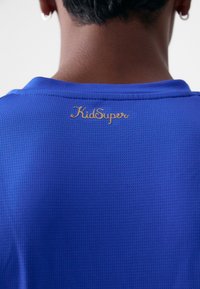 Modra športna majica iz teksturirane mrežaste tkanine, z zlatim logotipom "KidSuper" na zadnji strani blizu ovratnika.