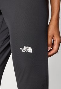 Crne atletske tajice napravljene od glatkog, rastezljivog materijala s bijelim logom "The North Face" na donjem dijelu noge.