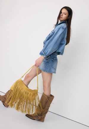 Jeune femme portant une veste en jean oversize, une jupe en jean et des bottes marron montantes, tenant un grand sac à main jaune à franges.