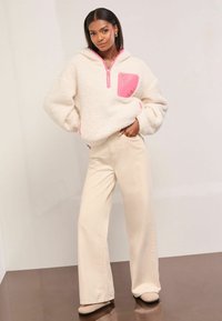 Pulover alb din Sherpa cu fermoar și buzunar roz; asociat cu pantaloni largi crem și pantofi slip-on bej. Textură moale, croială relaxată.