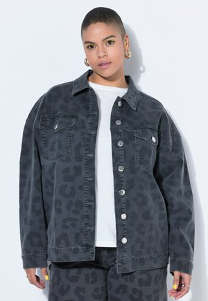 Ung person med kort hår iført mørk leopardprint denimjakke og bukser, hvid skjorte, guldfarvede rundøreringe og gul øjenskygge.