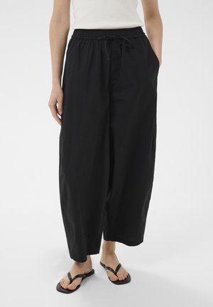 Femme portant un pantalon noir ample avec un cordon de serrage et des sandales noires, debout avec les mains détendues le long du corps devant un fond uni.