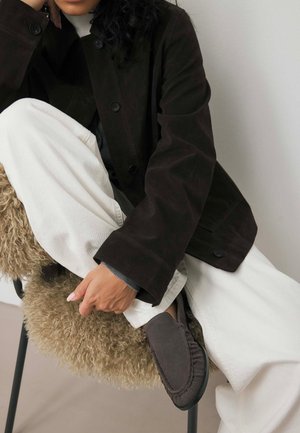 Veste en suédine marron foncé avec des boutons, associée à un pantalon large écru. La main repose sur une chaise en fausse fourrure beige et duveteuse, portant des chaussures grises.
