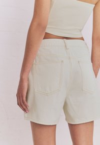 Shorts in denim beige chiaro con taglio dritto, dotati di tasche posteriori e cuciture decorative. Il tessuto ha una trama liscia.