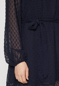 Temno modra obleka z teksturiranim krožnim vzorcem, prosojnimi rokavi in zavezanim pasom v pasu. Elastični manšeti z nabranim detajlom.