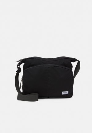Sac bandoulière en tissu noir avec une bandoulière réglable, une fermeture à glissière et une grande poche avant zippée, présenté sur un fond uni.