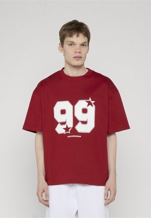 Młody mężczyzna w luźnej czerwonej koszulce z białymi grafikami "99" i gwiazdą, w białych spodniach, stojący na jednolitym jasnym tle.