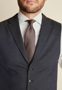 Chaleco gris oscuro con dos botones, combinado con una camisa a rayas en azul claro y blanco y una corbata marrón texturizada.