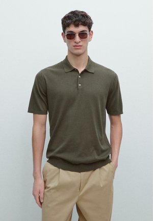 Olivově zelené polokošile s krátkými rukávy, tříknoflíkovým límcem a pletenou texturou. Nošena s béžovými kalhotami, model stojí na světlém pozadí.