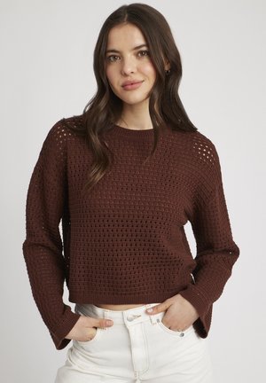 Pull en maille marron à coupe ample, avec un tissage ouvert texturé, des manches longues et des poignets côtelés, associé à un pantalon blanc taille haute.