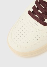 Gros plan sur l'avant d'une basket couleur crème avec des détails perforés et des lacets bordeaux foncé sur un fond blanc.