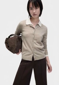 Béžový svetr na knoflíky s kontrastním bílým límečkem. Modelka drží hnědou koženou kabelku. Vzhled doplňují tmavé kalhoty.