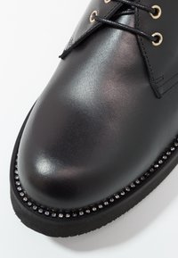 Scarpa in pelle nera con una texture liscia, punta arrotondata e occhielli dorati. Presenta una fila decorativa di strass trasparenti lungo la suola.
