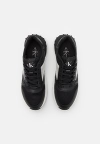 Černé sportovní boty se síťovaným svrškem, bílými proužky, kulatou špičkou, texturovanou podrážkou a viditelným šněrovacím systémem. Logo značky na jazyku.