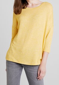 Żółty, lekki sweter wykonany z dzianiny o luźnym kroju, szerokim dekolcie i rękawach trzy czwarte, zestawiony z szarymi dżinsami o slim fit.