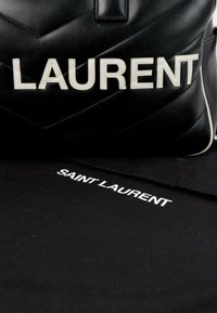 Sac en cuir matelassé noir avec de grandes lettres blanches cousues formant le mot LAURENT, et sac anti-poussière noir avec texte blanc SAINT LAURENT.