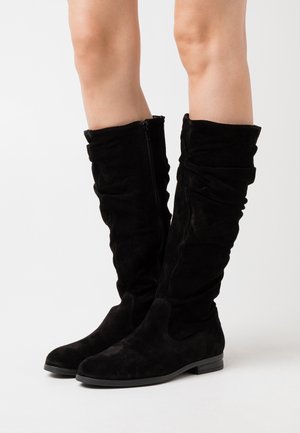 Nogi w czarnych zamszowych kozakach sięgających do kolan, z luźno opadającymi cholewkami i niskimi płaskimi obcasami, stojące na jasnym, jednolitym tle.