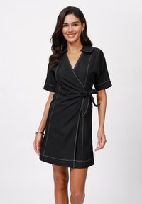 Black wrap dress with short sleeves, featuring white stitching along the neckline, waist, and hem, finished with a tied belt detail.