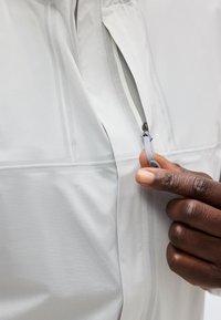 Χέρι τραβάει ένα γκρι φερμουάρ στην τσέπη ενός λευκού μπουφάν, δείχνοντας υφή υφάσματος και κοντινό πλάνο δαχτύλων.