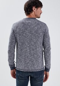 Camicia grigia a maniche lunghe con texture e scollo rotondo, con una finitura grigia più scura al colletto e ai polsini, abbinata a jeans blu scuri.