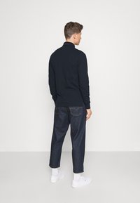 Polo shirt bleu marine à manches longues avec un col, associé à un jean tapered bleu foncé. Baskets blanches et ligne de chaussettes visible. Fond uni.
