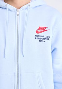 Világoskék zipzáras kapucnis pulcsi húzózsinórral; a piros Nike logóval és tengerészkék "CSAK KÉSZLETES SZEMÉLYEKNEK" felirattal rendelkezik. Puha szövet textúrával.