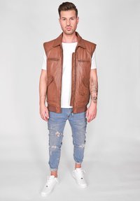 Brun læder ærmeløs vest med forlommer, båret over en hvid t-shirt. Kombineret med slidte lyseblå jeans og hvide sneakers.