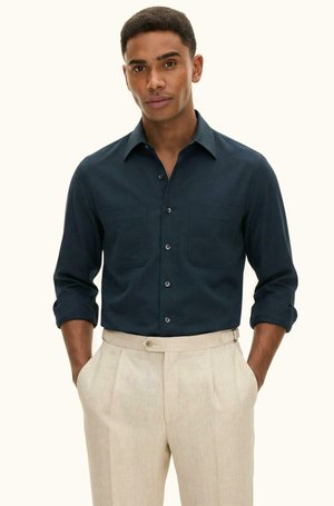 Mann trägt ein dunkelblaues Hemd mit Knöpfen und hochgekrempelten Ärmeln sowie beige hochtaillierte Hosen und steht mit den Händen in den Taschen.