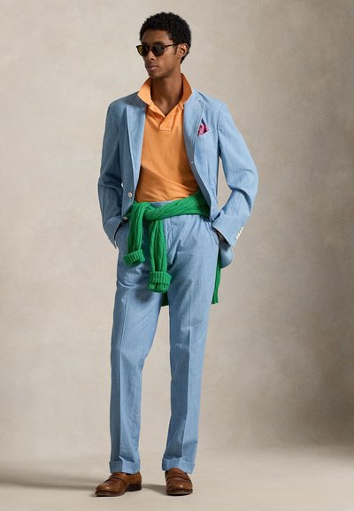 Σακάκι ανοιχτού γαλάζιου χρώματος με ταιριαστό παντελόνι, πορτοκαλί πόλο, πράσινο πουλόβερ δεμένο γύρω από τη μέση, καφέ παπούτσια και γυαλιά ηλίου, με ένα μαντιλάκι στην τσέπη.