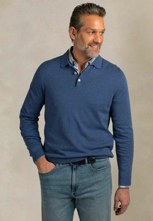 Mann mit grauem Bart, der einen blauen Pullover über einem Hemd mit Kragen und blaue Jeans trägt, mit einer Hand in der Tasche steht, nach rechts schaut und lächelt.