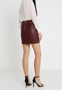 Femme portant un chemisier blanc à manches longues, une mini-jupe en cuir marron avec fermeture éclair au dos, et des sandales noires à talons hauts avec bride à la cheville, debout.