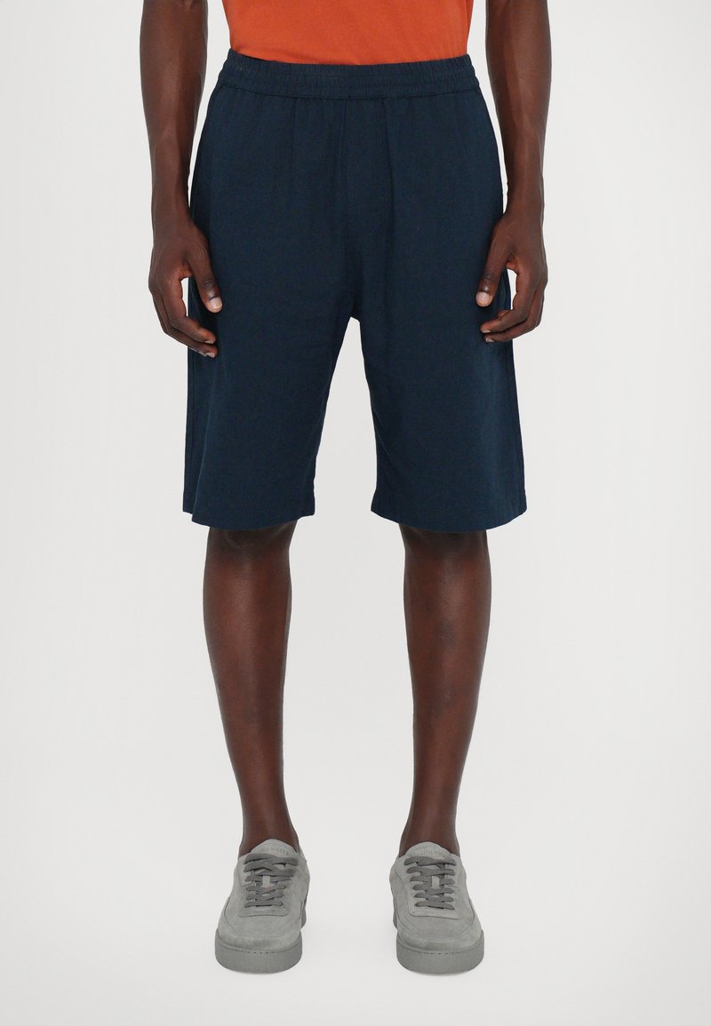 Shorts bleu marine en tissu léger, dotés d'une taille élastique et de poches latérales, tombant au-dessus des genoux, associés à des chaussures grises.