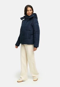 Navy blue puffer jacket with a hood, zip closure, multiple pockets, and adjustable drawstring. Paired with cream wide-leg trousers and light shoes.