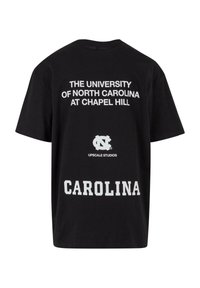 Mustat puuvillapaidat, joissa valkoiset tekstit: "NORTH CAROLINA YLIOPISTO CHAPEL HILLISSA" ja "CAROLINA", sekä "UPSCALE STUDIOS" logo.