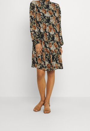 Kvinde iført en sort knælængde kjole med blomsterprint, lange ærmer og brune hælede sandaler, stående mod en ensartet lys baggrund.