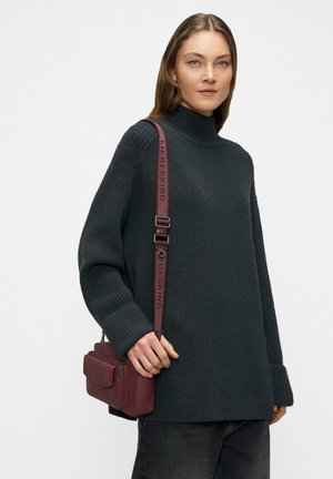 Kobieta o prostych brązowych włosach nosi czarny, teksturowany golf i ma małą bordową torebkę na ramię z pasekiem z logo marki.