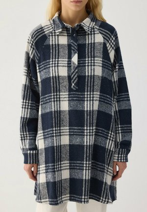Un mannequin porte une longue robe-chemise à carreaux bleu marine et blanc, avec un col boutonné et des manches longues.