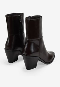 Botas de tobillo de cuero marrón con punta afinada, cierre de cremallera y tacones cónicos apilados. Textura suave con sutiles detalles de costura.