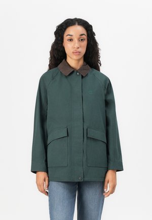 Vrouw met lang krullend haar, gekleed in een donkergroene jas met een bruine kraag en voorflapzakken, gecombineerd met blauwe jeans, tegen een witte achtergrond.