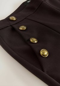 Detailní záběr černé látky se čtyřmi zdobenými zlatými knoflíky uspořádanými ve dvou vertikálních řadách na panelu oděvu.