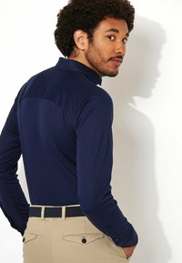 Marineblauwe polo met lange mouwen en een kraag, met een gladde textuur en een getailleerd ontwerp, gecombineerd met beige broek en een donkere riem.