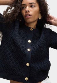 Donna con capelli ricci che indossa un cardigan navy testurizzato con grandi bottoni dorati e orecchini ovali dorati abbinati, in posa con le braccia sollevate.
