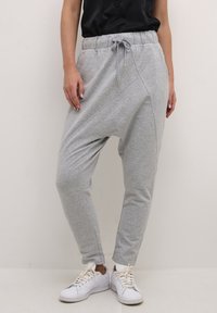 Vrouw in lichtgrijze drop-crotch sweatpants met elastische tailleband en trekkoord, gecombineerd met witte sneakers en een zwart topje.