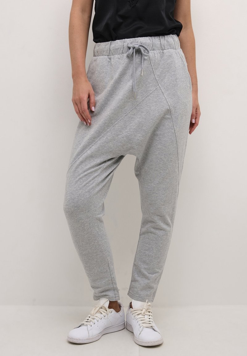 Vrouw in lichtgrijze drop-crotch sweatpants met elastische tailleband en trekkoord, gecombineerd met witte sneakers en een zwart topje.